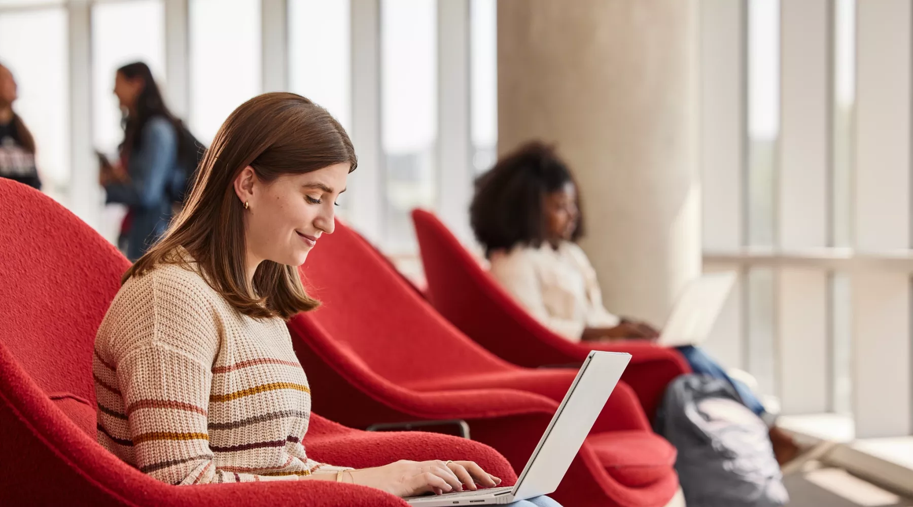 Lexi Raikes sitting on the second floor of the Belknap Academic Building, in a red chair, typing happily on her laptop