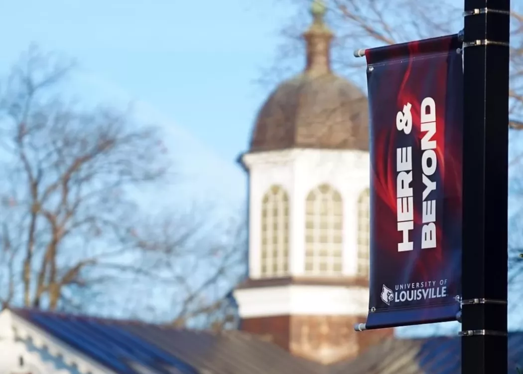 UofL's law school copula is out of focus in the background framed byt a lamppost banner reading "Here & Beyond" with UofL branding.